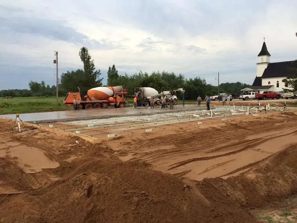 Construction site with concrete mixers on a muddy road near a white church tower under a cloudy sky