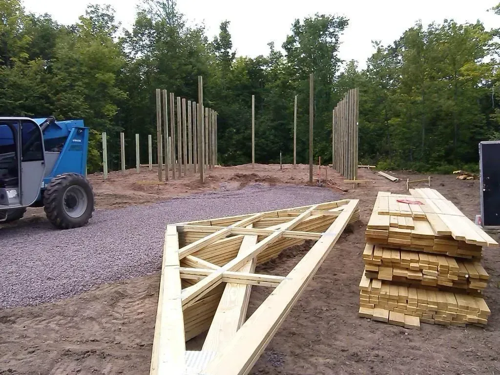 Construction site with stacked lumber, wooden frame pieces, gravel, and a blue tractor near a tree line