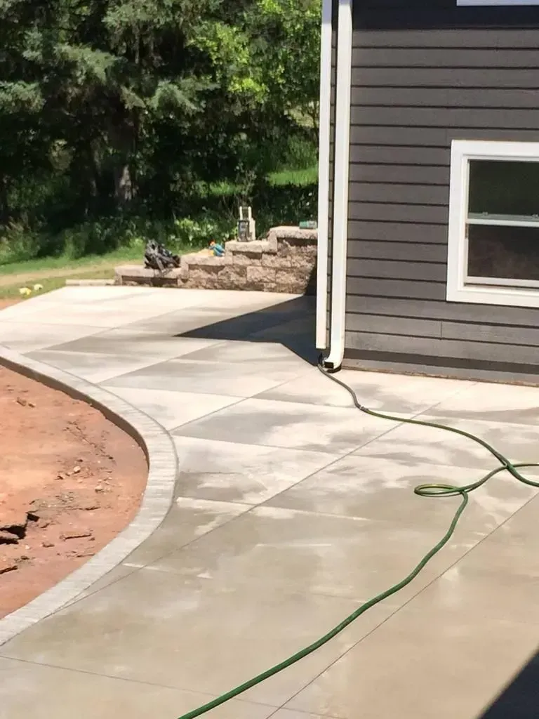 Curved concrete walkway beside a gray house, with a green garden hose across the pavement.
