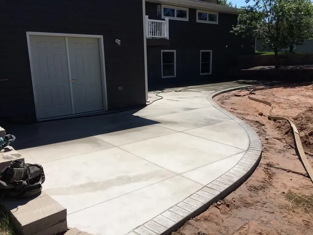Curved concrete walkway beside a dark building, with a dirt landscaping area on the right.