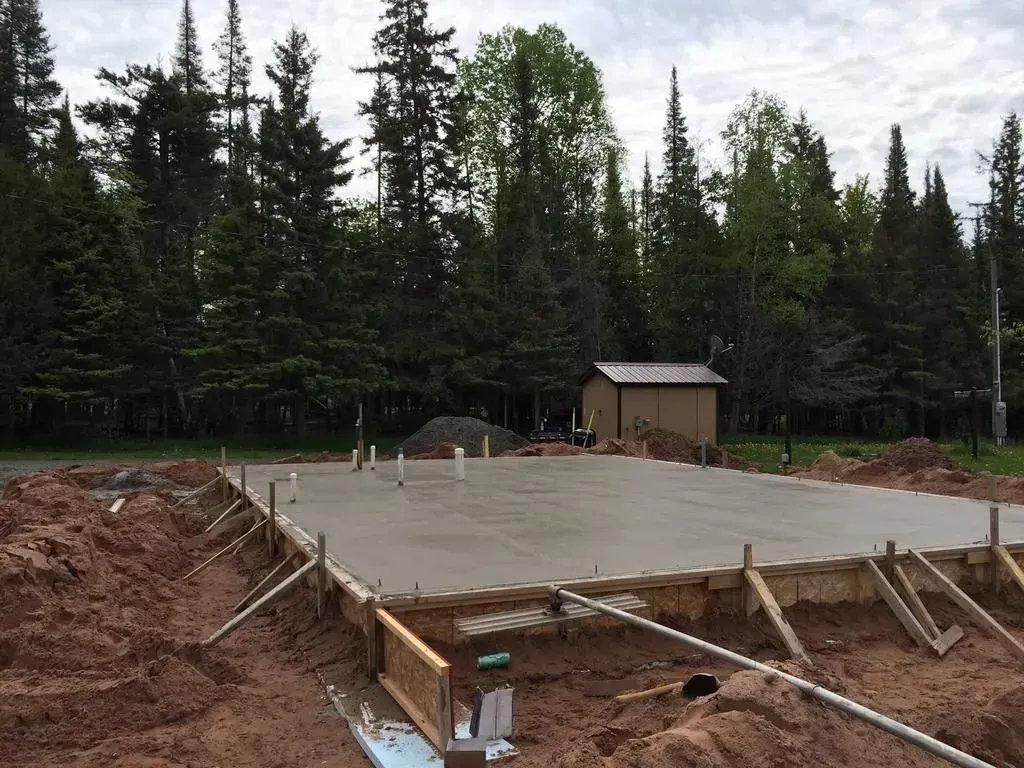 Concrete foundation under construction in a forest clearing, with lumber forms and a small shed nearby.