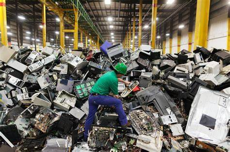 Worker in a green shirt and blue pants amidst a mountain of discarded electronics in a recycling facility.