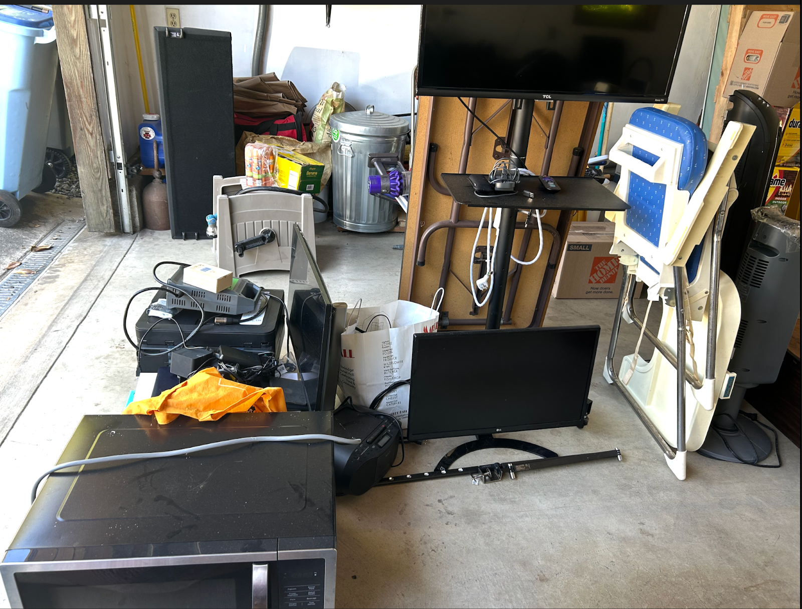 Cluttered garage with a microwave, TV, printer, high chair, trash can, and other household items.