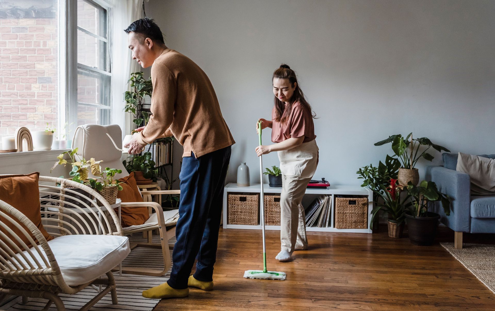 Couple tidying their living room, one sweeping the floor while the other works at a desk