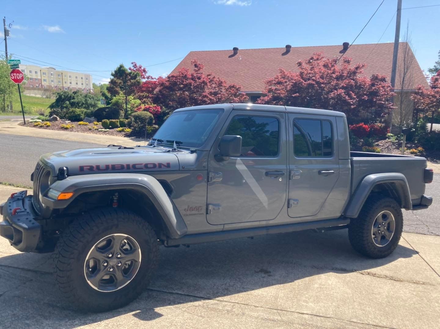 Gray Jeep Gladiator Rubicon parked on a paved road, near a building and blooming trees.
