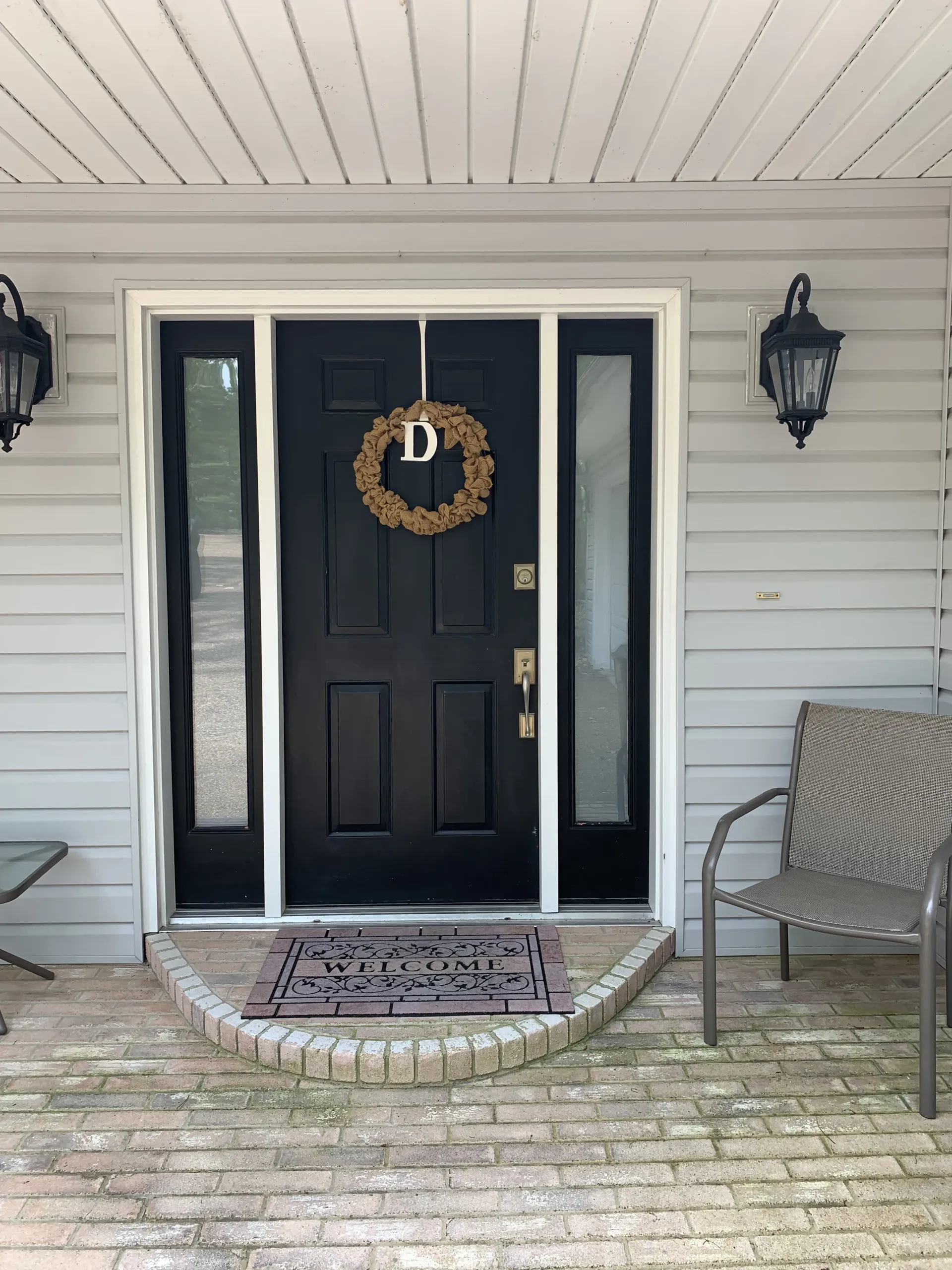 Black front door with wreath and sidelights, flanked by sconces. A welcome mat rests on a brick porch.