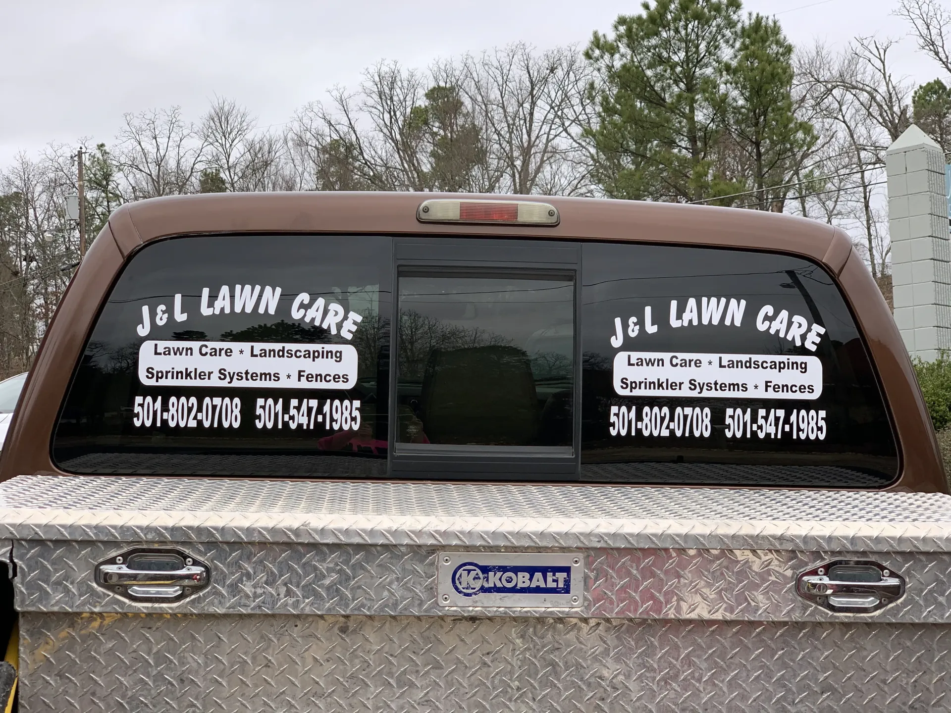 Brown pickup truck with J & L Lawn Care signage on the rear window and a metal tool box.