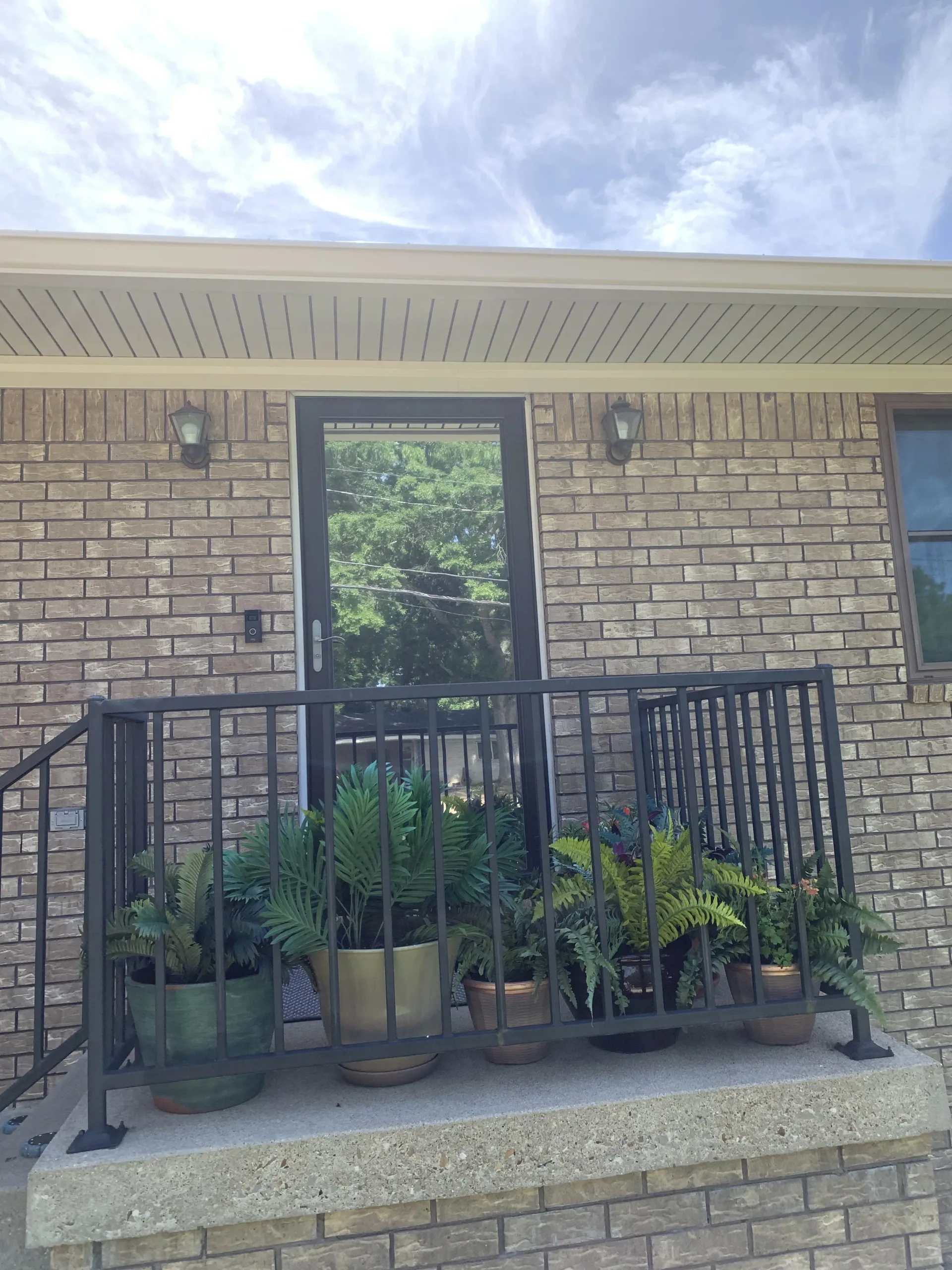 Brick building entrance with a black screen door, black railing, and potted plants.