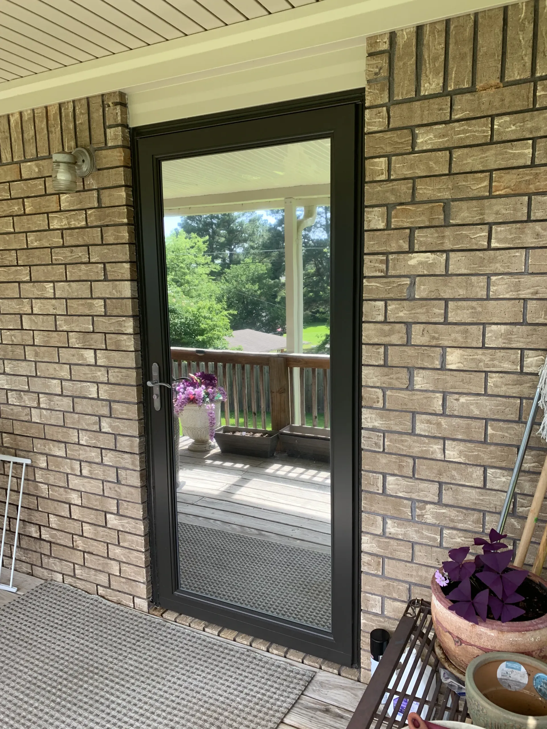 Black framed screen door on a brick wall, reflecting a deck with potted plants and trees.