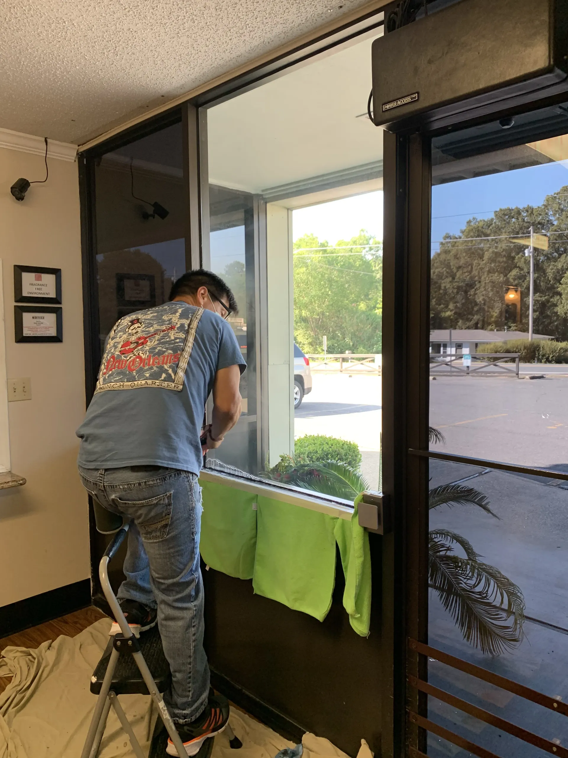 A person on a step stool installs window film. A green towel and light shines through window.