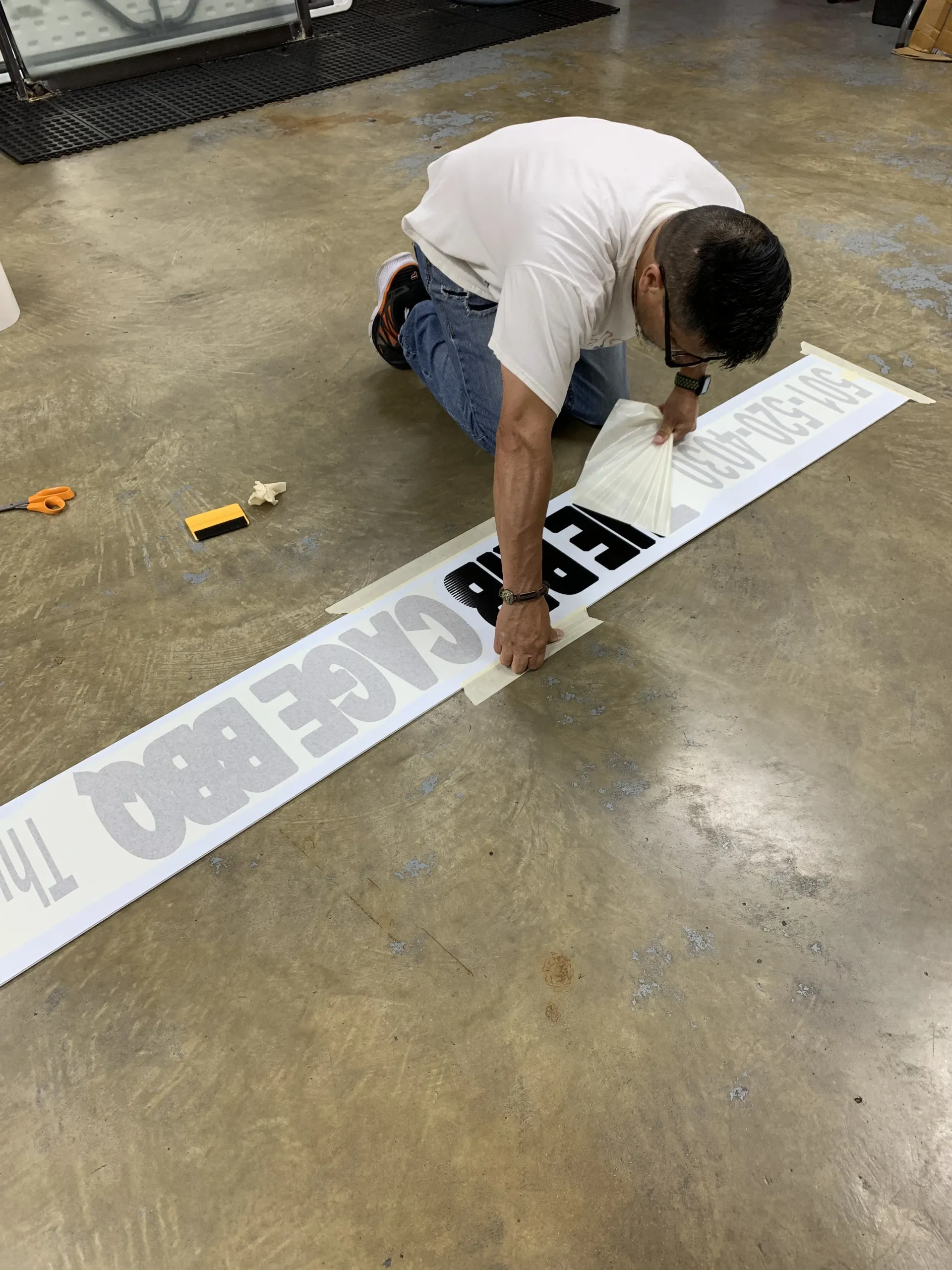 Person kneeling, applying large lettering to a surface on the floor; text says