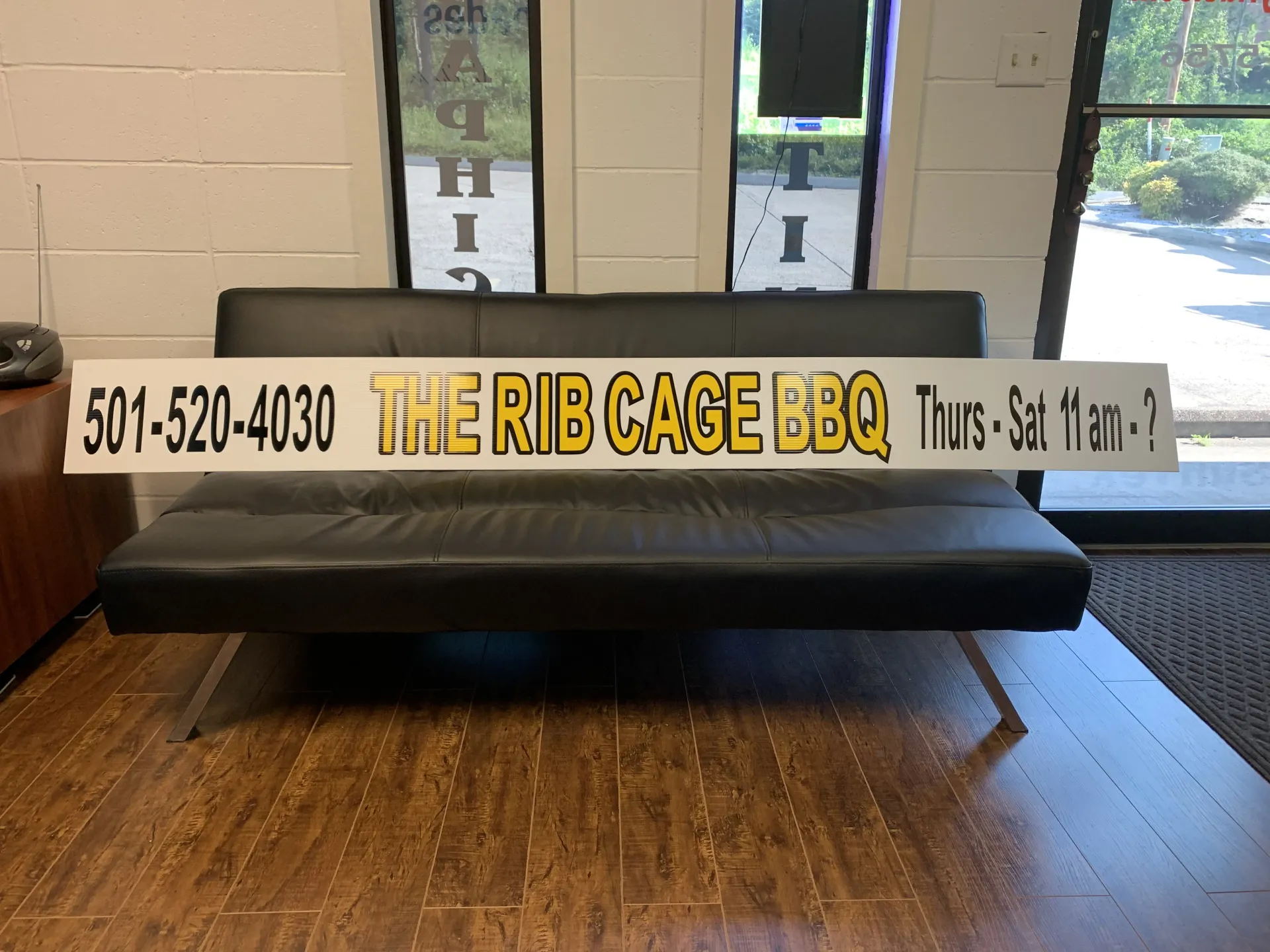 Sign for The Rib Cage BBQ restaurant, including a phone number, hours, and logo, displayed on a black couch.