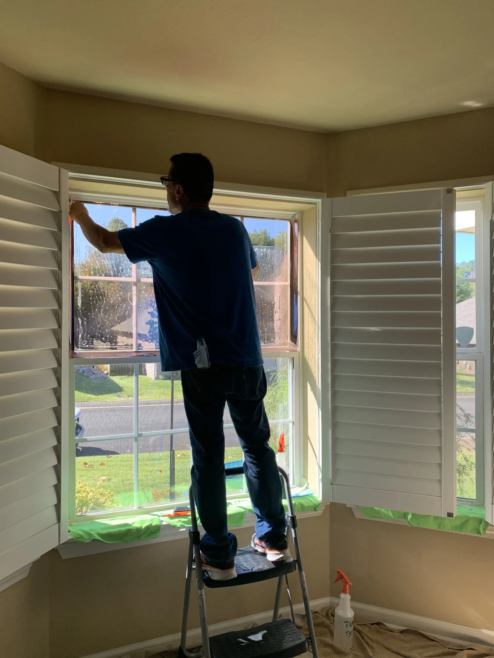 Person standing on a step stool cleaning a window inside a house with white shutters.