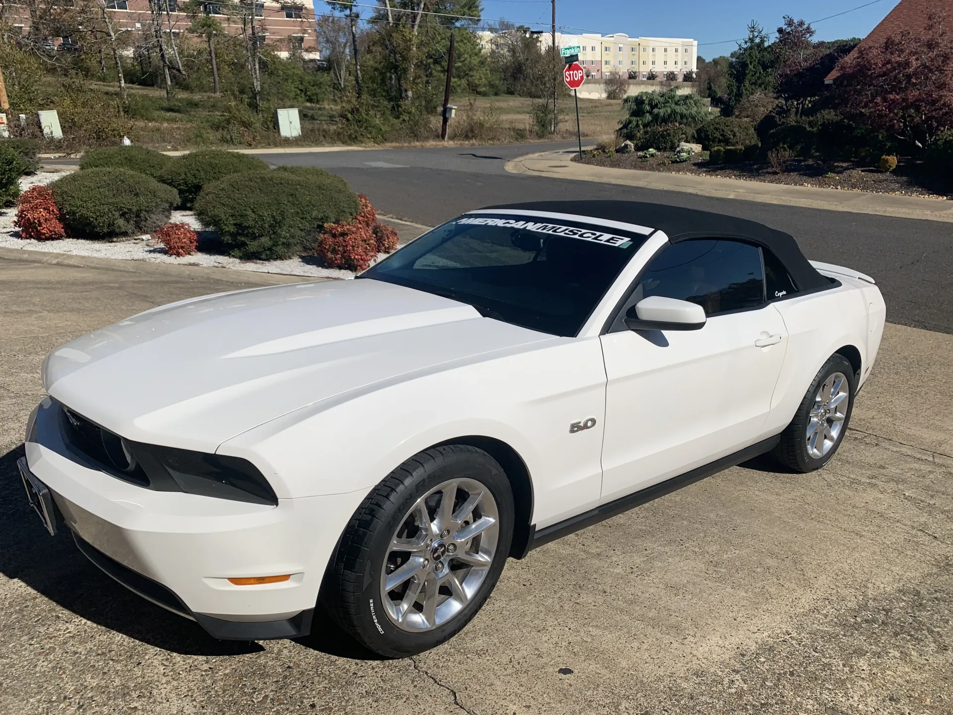 White convertible Ford Mustang parked outdoors. Black top, chrome wheels, and sunlight.