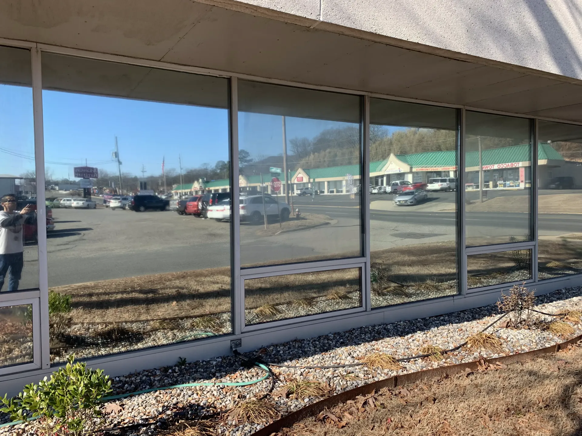 Exterior of building with large windows reflecting a parking lot and businesses on a sunny day.