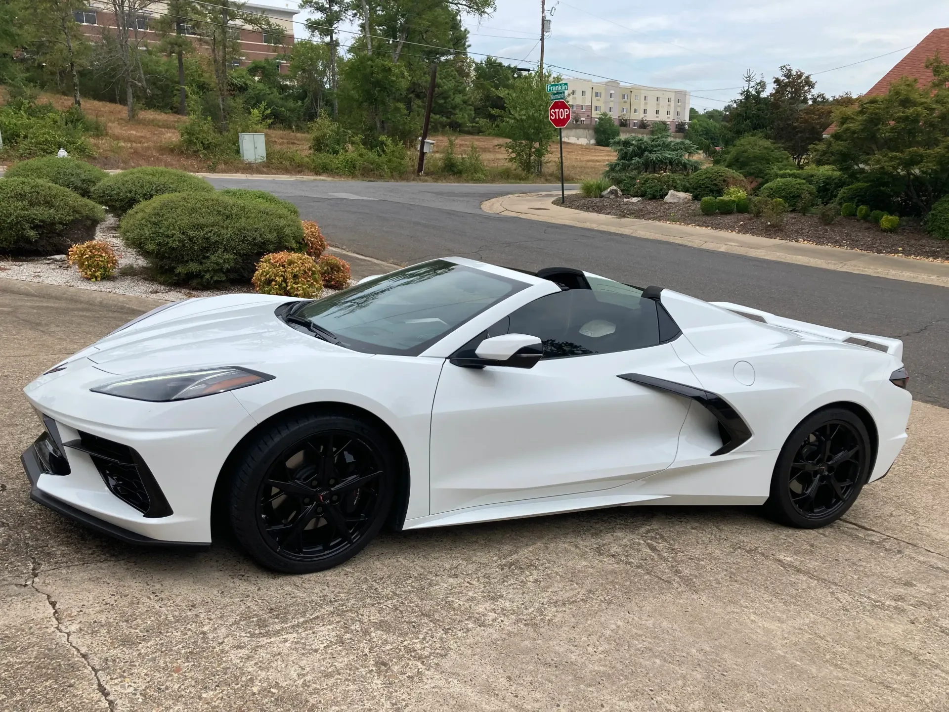 White convertible sports car with black wheels parked on a driveway near green bushes and a building.