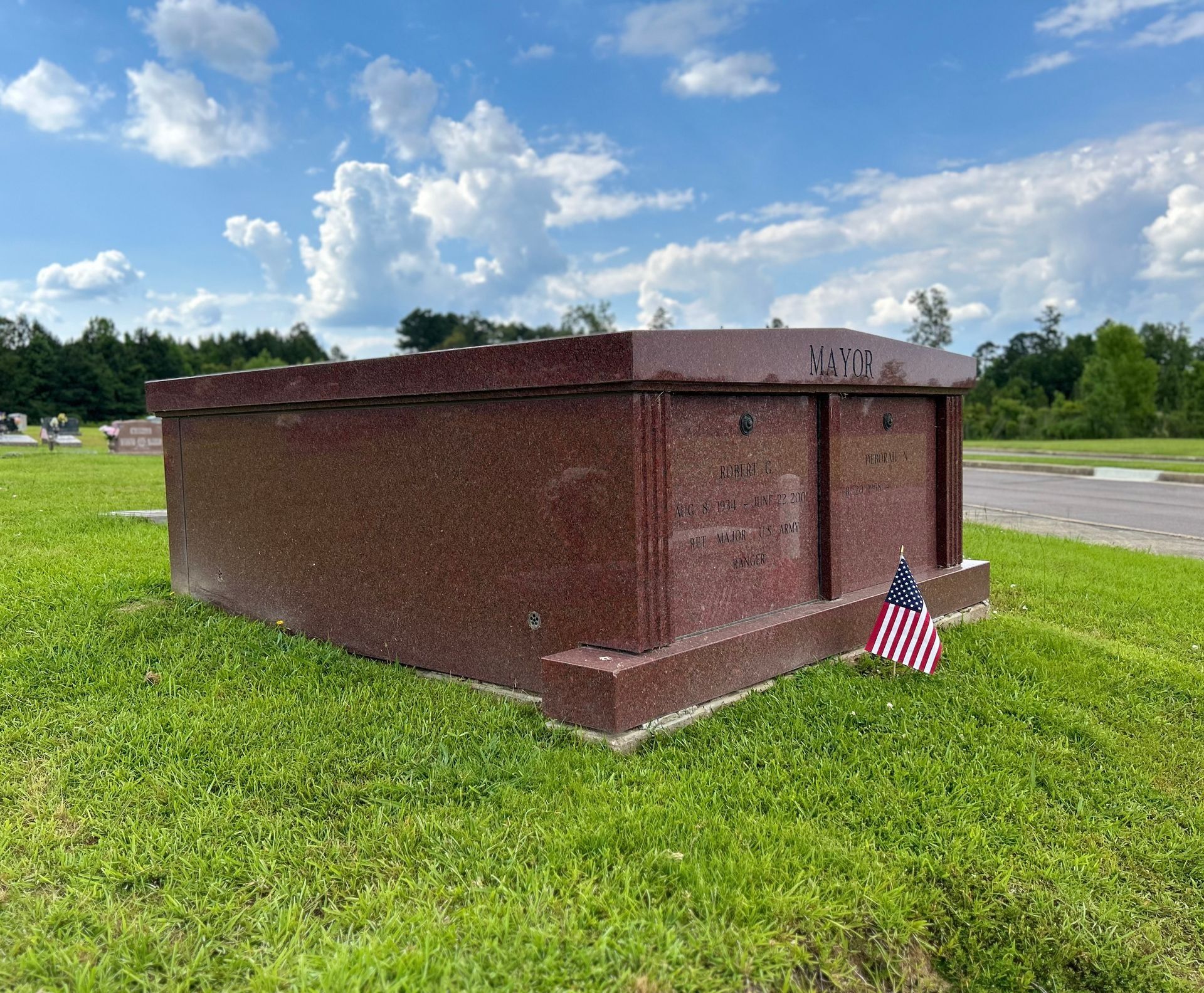 Red granite memorial tomb in a grassy cemetery with a small American flag beside it under a cloudy sky
