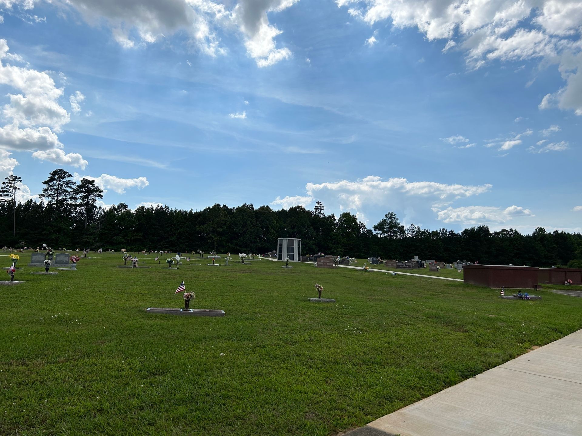 Wide grassy park with scattered people, a concrete path, trees, and a partly cloudy blue sky