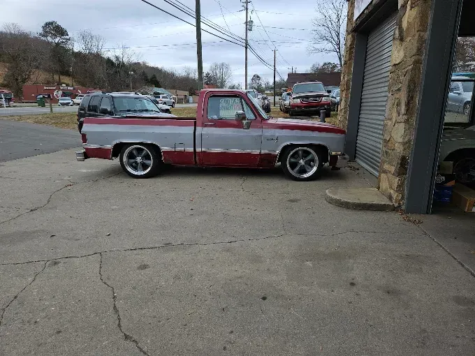 Red and white lowered pickup truck parked beside a garage on a cracked roadside.