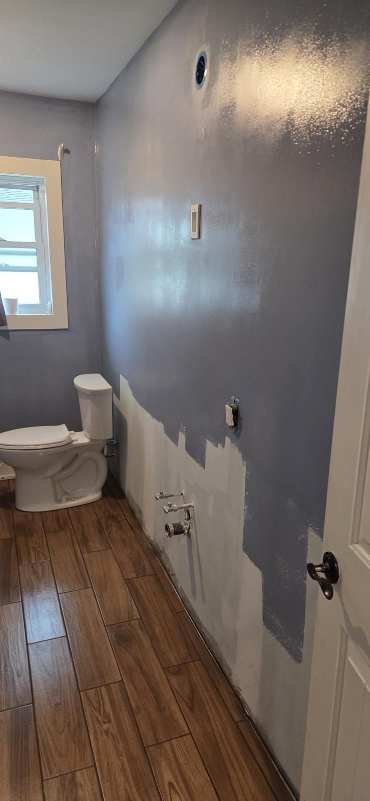Small bathroom with gray walls, wood floor, toilet, window, and exposed plumbing on the wall.