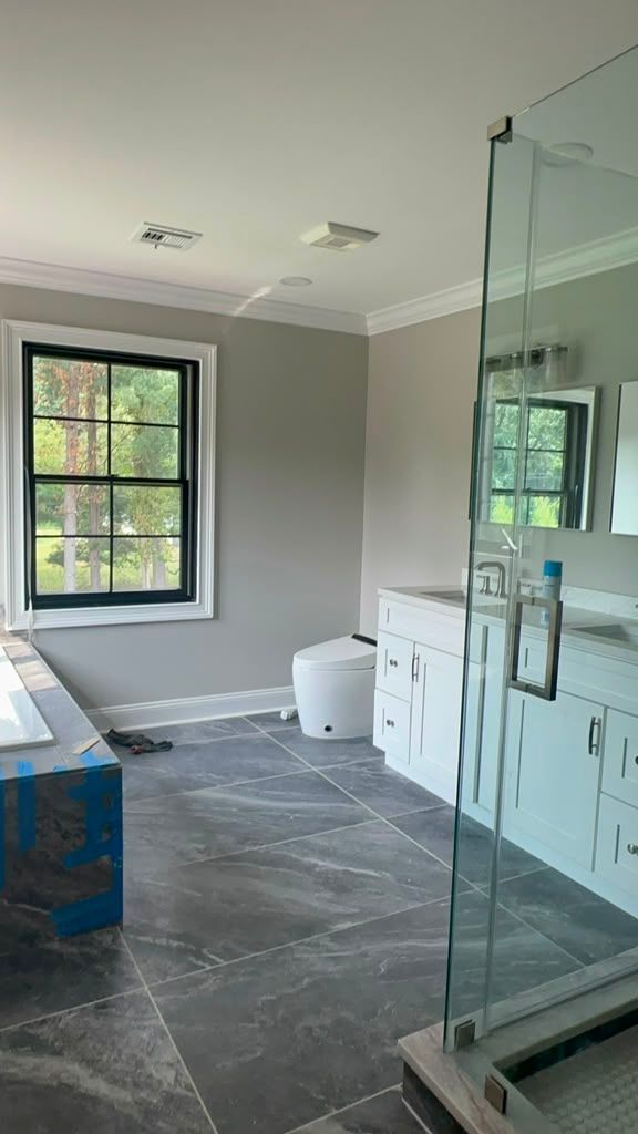 Modern bathroom under construction with gray tile, window, vanity, toilet, and glass shower enclosure