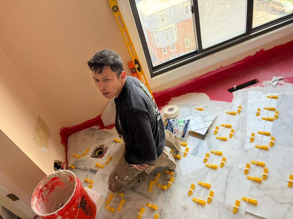 A person kneeling on a tiled floor during a renovation, installing marble tiles with yellow plastic spacers.