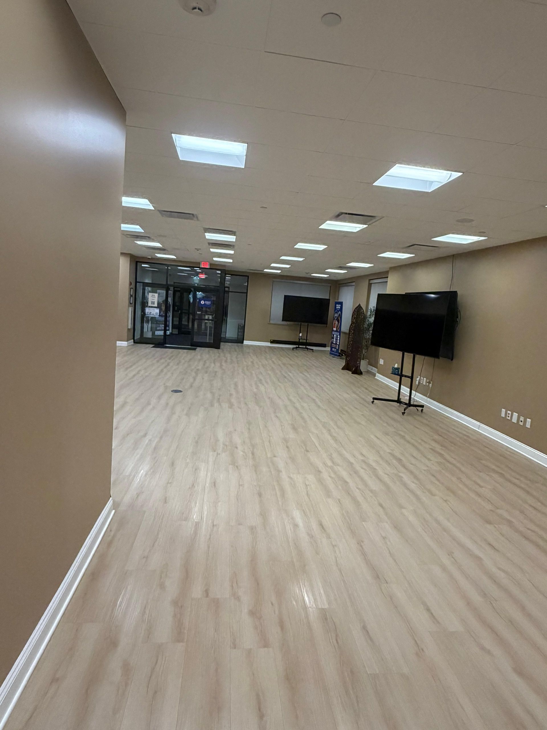 A large, empty room with light wood flooring, tan walls, and recessed lighting, featuring two rolling television stands.