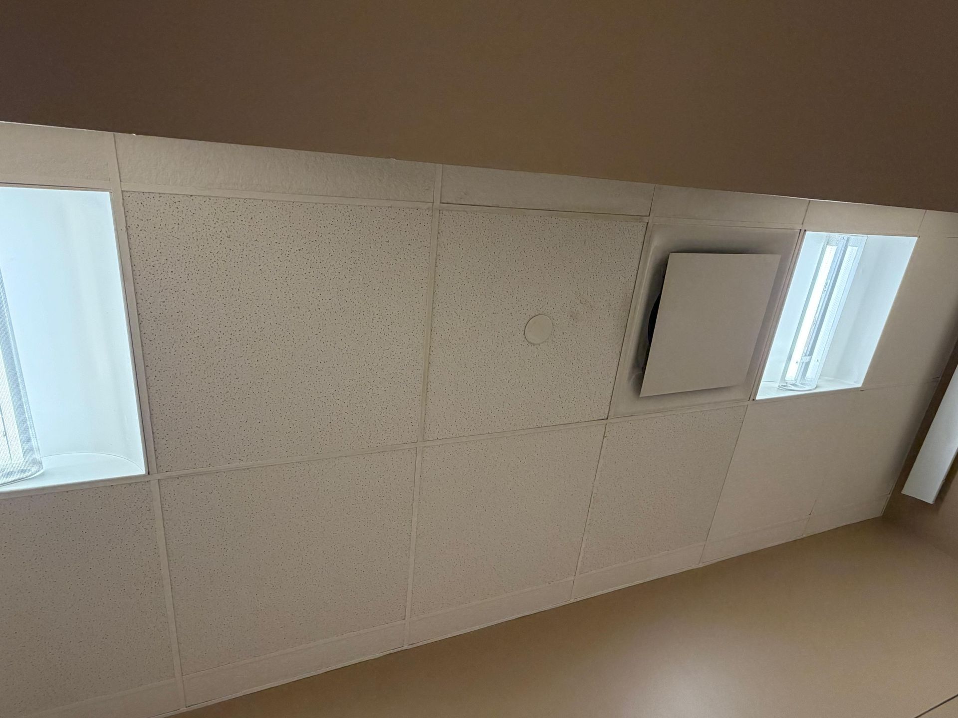 A white drop ceiling featuring textured square tiles, two recessed lighting fixtures, and a square access panel.