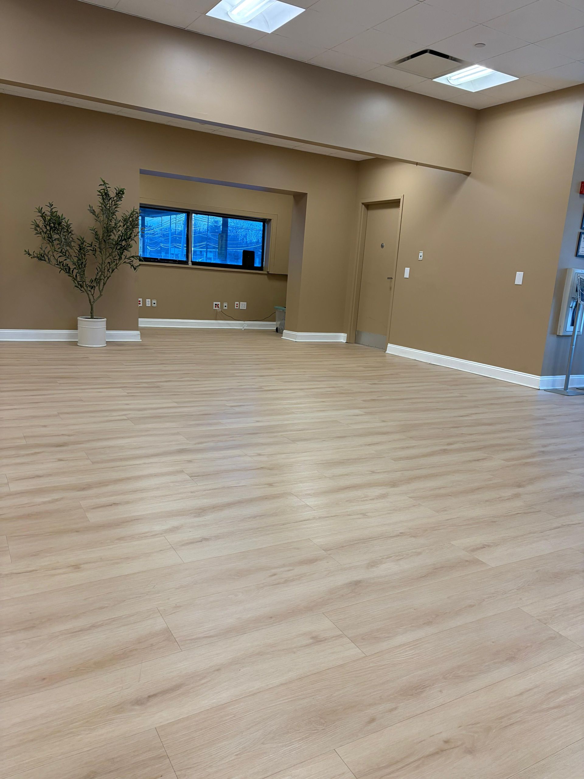 A spacious, empty room with light wood-look floors, tan walls, and an open doorway leading to a windowed area.