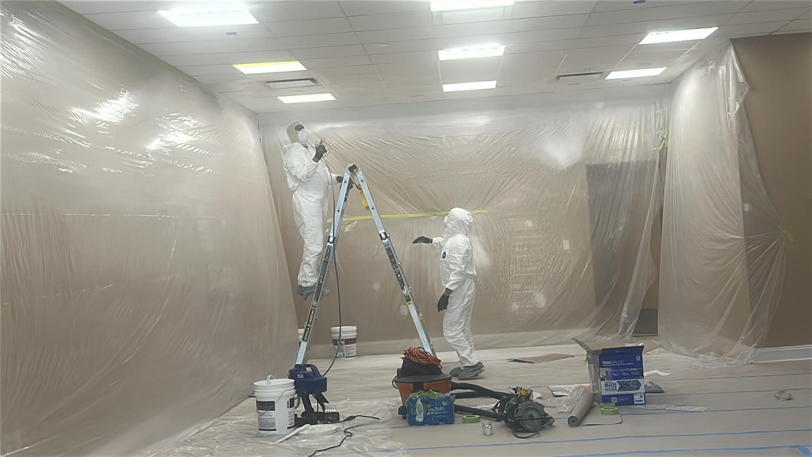 Two people in protective white suits work inside a room draped in plastic sheeting, one standing on a ladder.