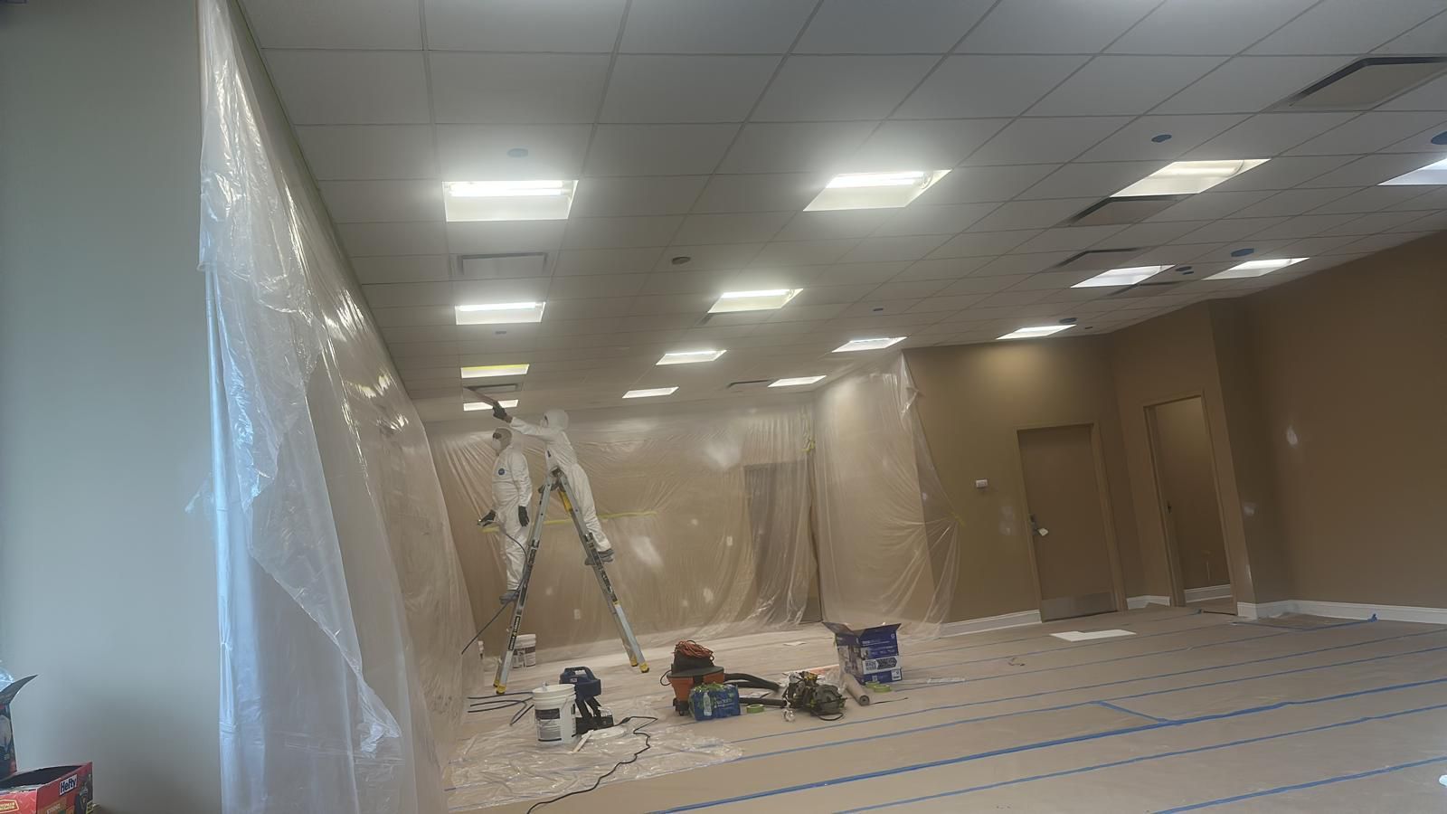 A commercial room under renovation with walls and floors covered in plastic sheeting, a ladder, and painting supplies.