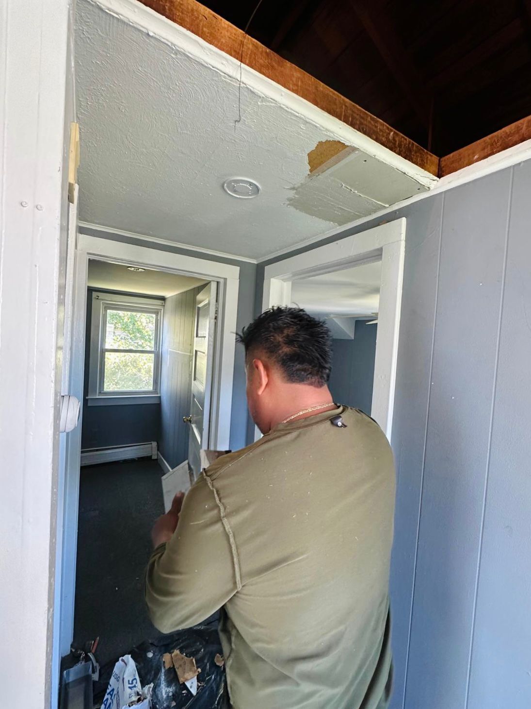 A person in a green long-sleeved shirt works on ceiling repairs in a gray-walled hallway with an open entryway above.