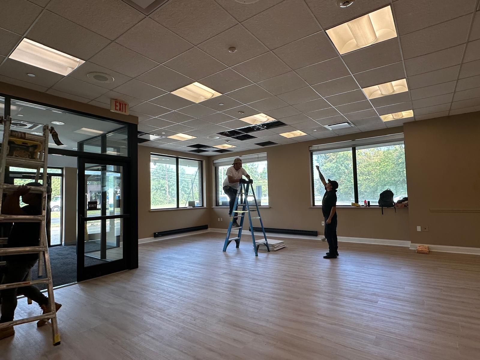 Workers perform maintenance on a ceiling in a modern office space with large windows and light wood-look flooring.