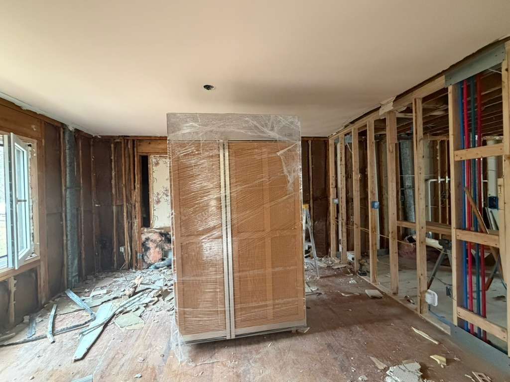 A large, wrapped stainless steel refrigerator stands in a room undergoing a home renovation with exposed wall studs.