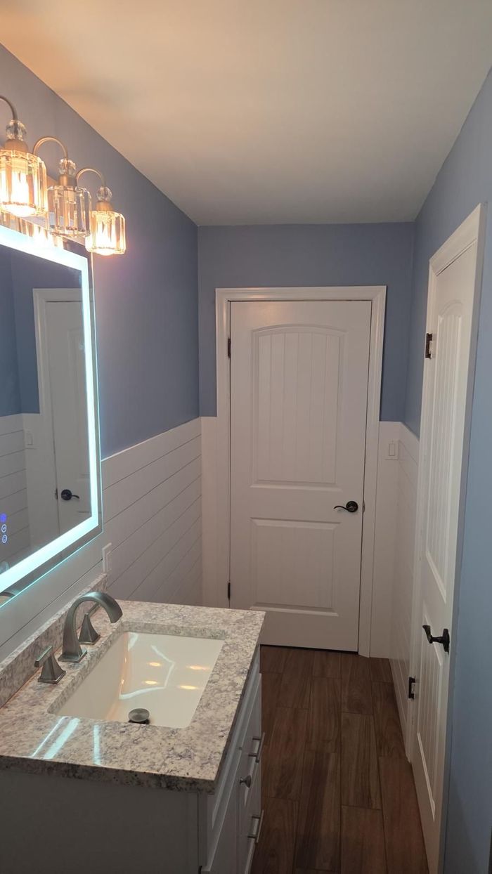 Narrow bathroom with blue walls, white vanity and door, lit mirror, and dark wood floor.