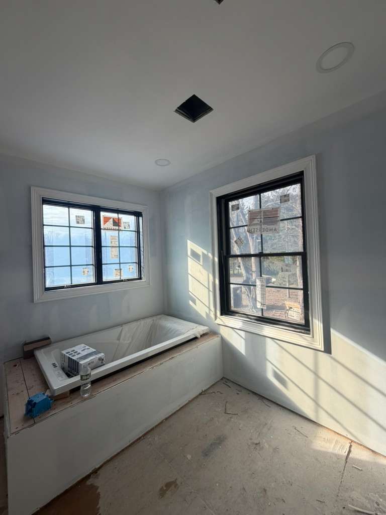 A bathroom under construction featuring a built-in white bathtub, two windows with black frames, and grey walls.