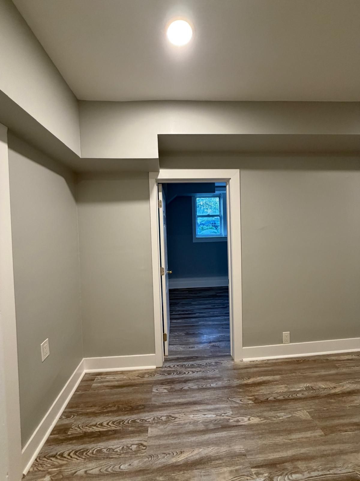Empty gray room with wood-pattern flooring and a doorway leading to a darker room with a window.