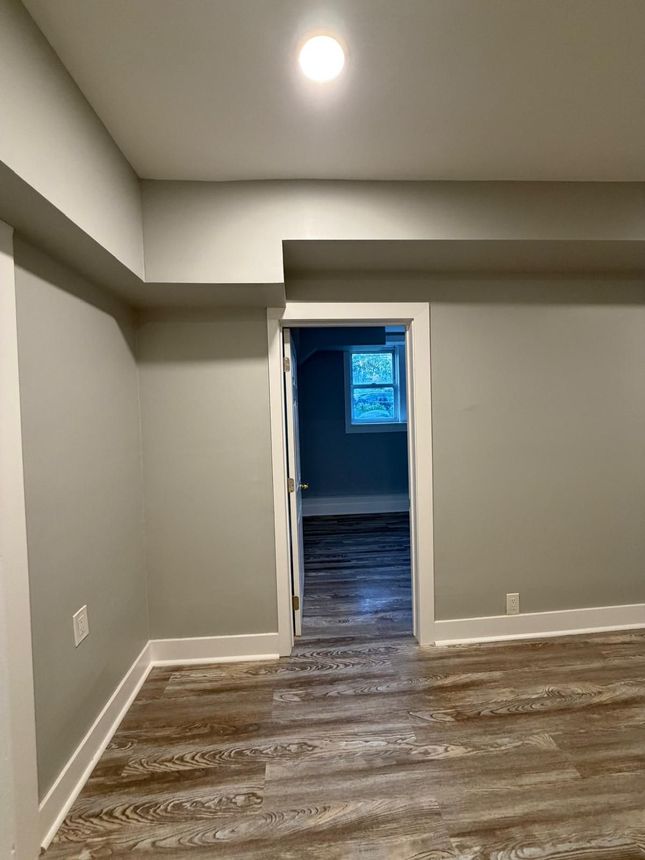 Empty gray room with wood-pattern flooring and a doorway leading to a darker room with a window.