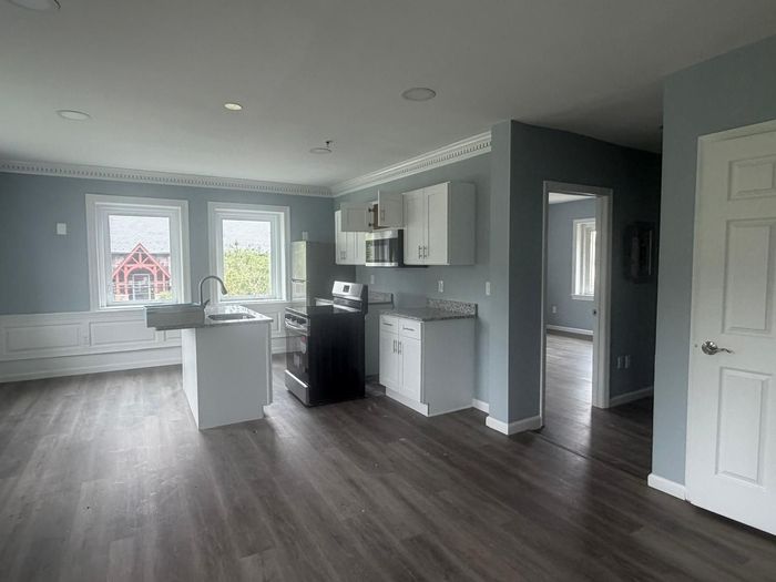Empty modern kitchen and living area with gray walls, white cabinets, and dark wood floors