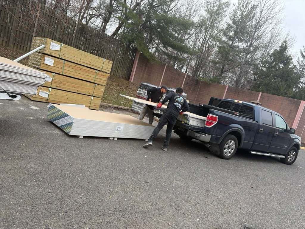 Two people load lumber from a stack onto the back of a black pickup truck parked in an outdoor lot.