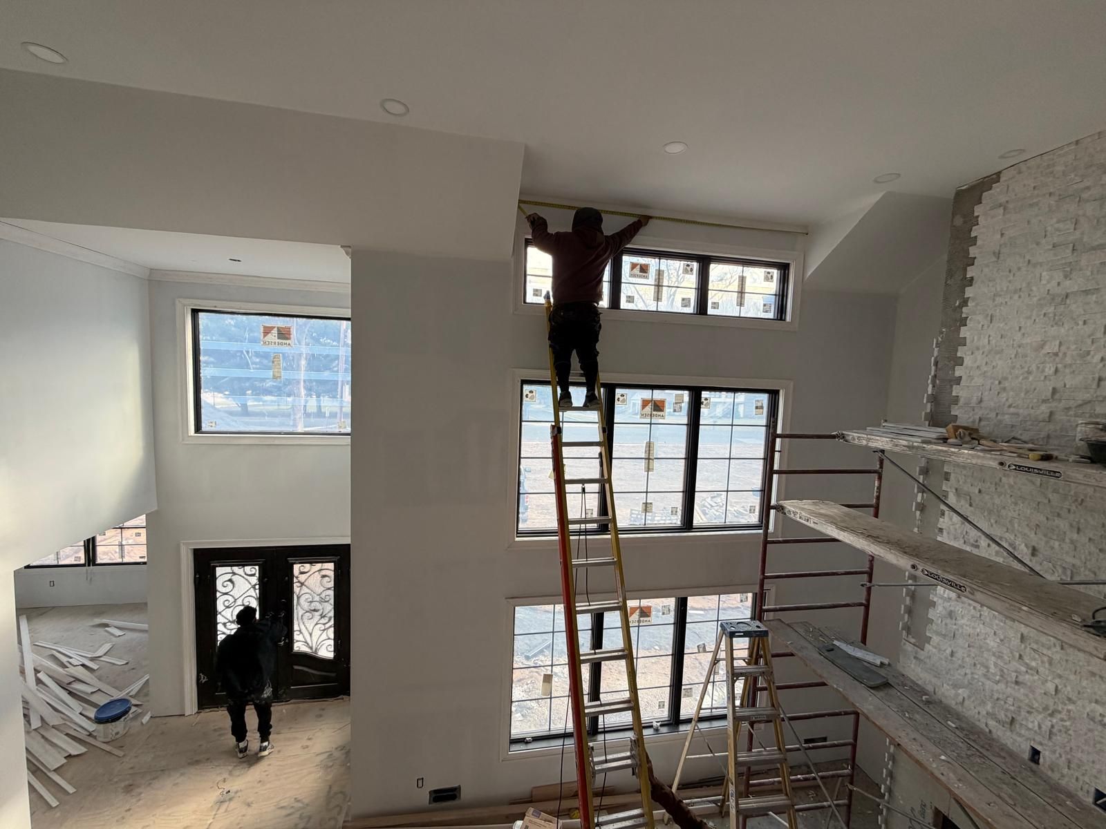 Workers on ladders in a bright, unfinished room with large windows and exposed brick.