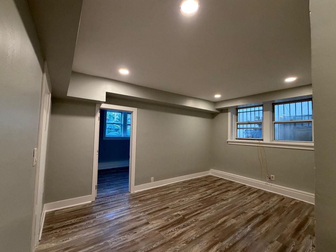 Empty gray living room with wood-patterned floor, recessed lights, and a doorway to a blue-lit room