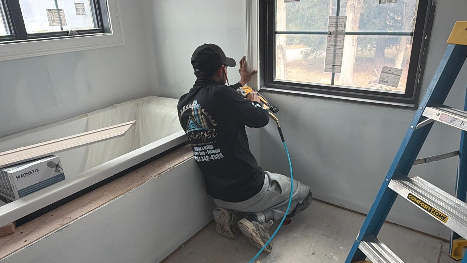 Person in black clothing spray-paints a wall in a bright room beside a window and ladder.