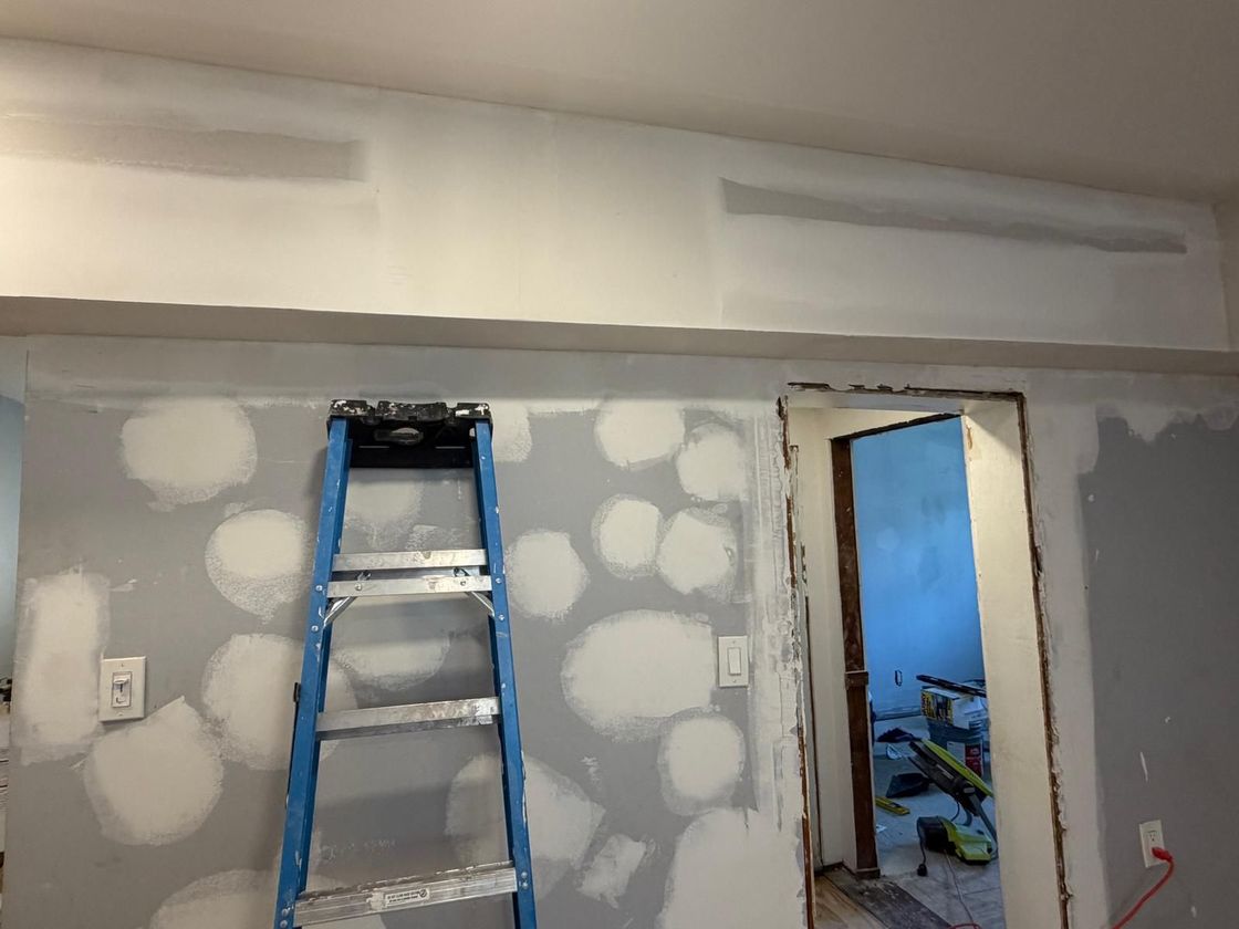 Gray drywall room under renovation with a blue ladder and an open doorway to a blue-lit adjacent room