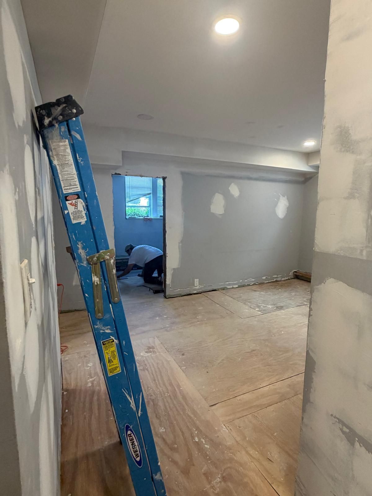 Renovation room with blue ladder, unfinished gray walls, plywood floor, and doorway to another room