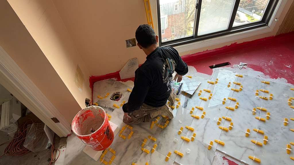 A worker kneels on a marble-tiled floor, using yellow tile leveling clips to install tiles in a room.