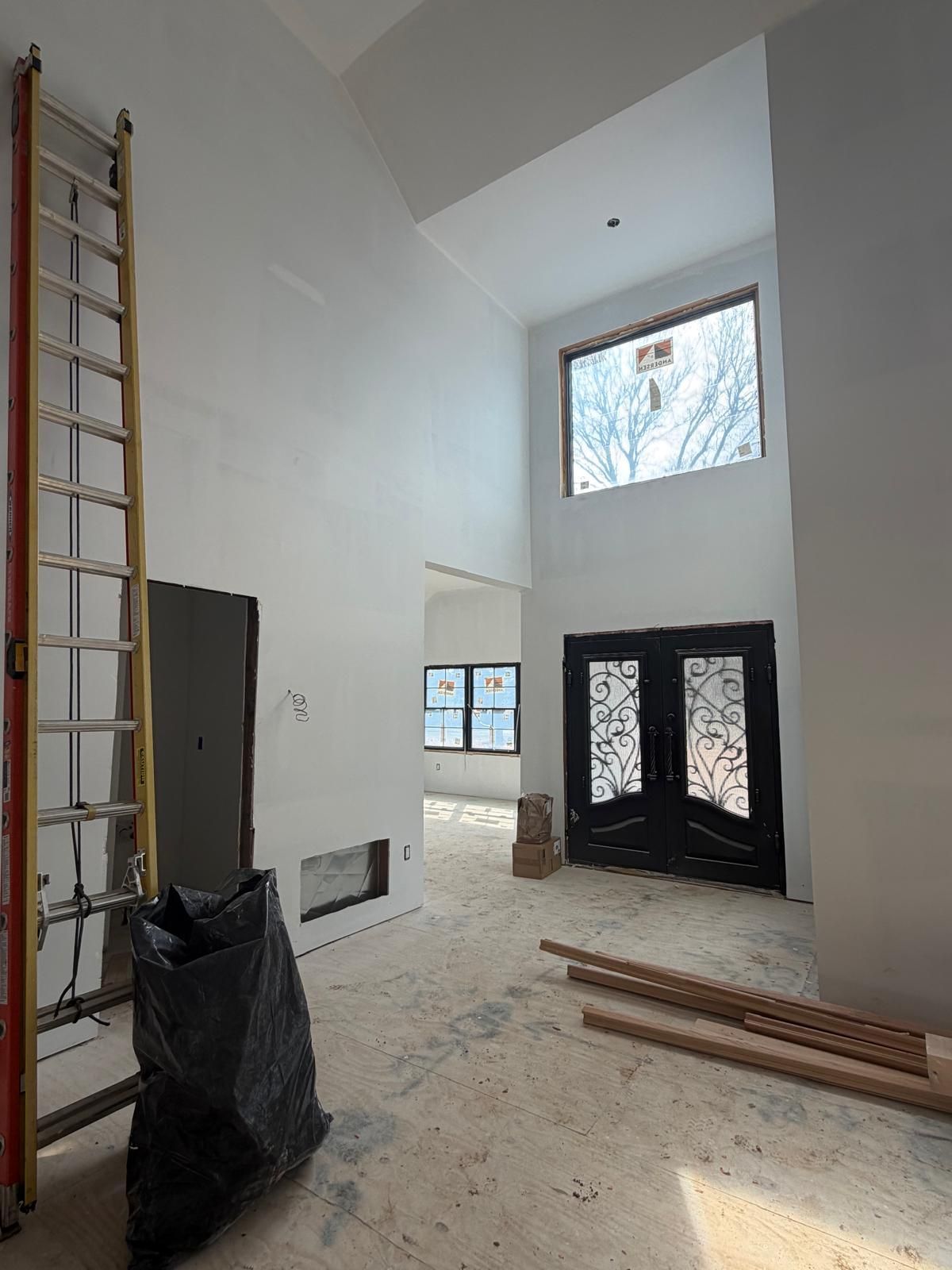 Interior of a bright unfinished room with high ceiling, ladder, black doors, and construction materials on the floor