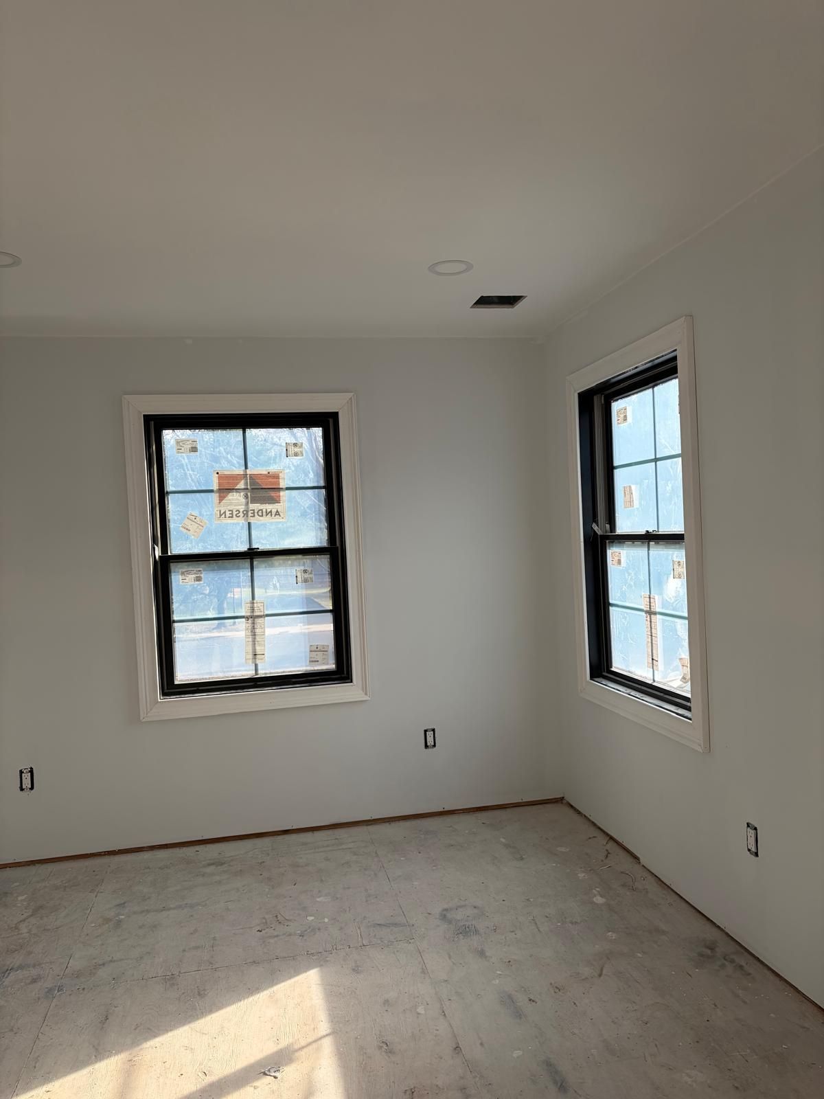 Empty white room with two windows and unfinished floors, lit by sunlight