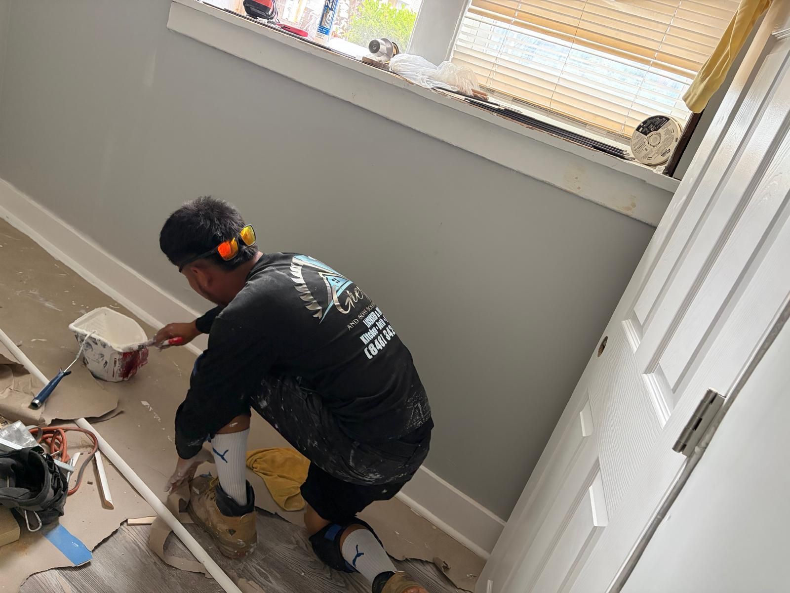 Worker painting a room wall near a window, crouched on a drop cloth with tools nearby.