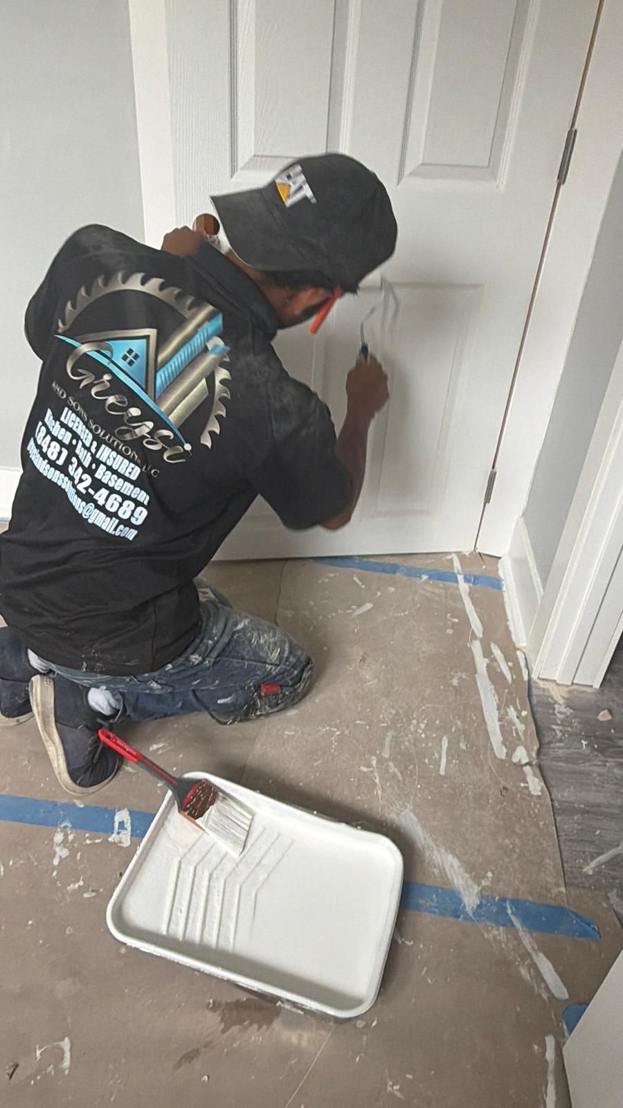 Worker painting a white door frame with a red brush in a hallway, paint tray on the floor.